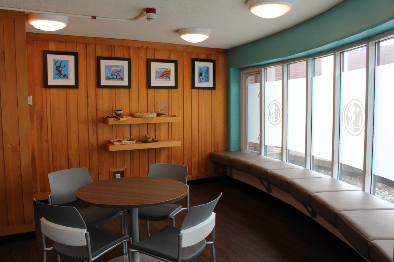 Mashkikì Pakesàyà Medicine Room. There is a decorated wooden wall at the back and a large curved window on the side with multiple seating spaces in the center of the room.