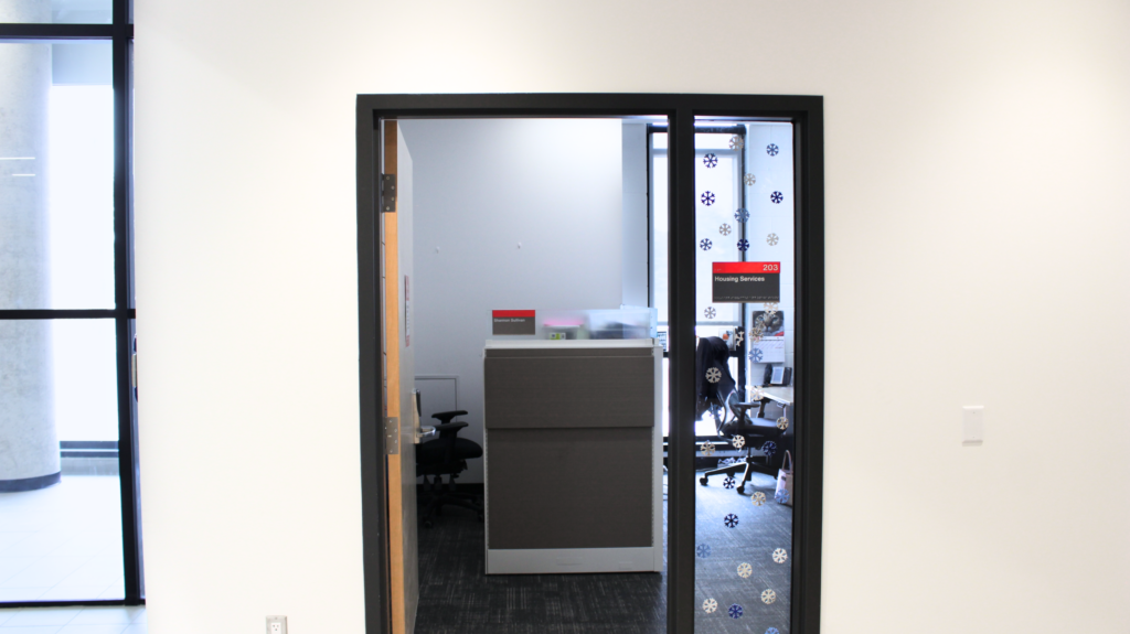 Head on shot of the open door to the Housing Services office with the room plaque on the right saying, "203 Housing Services"