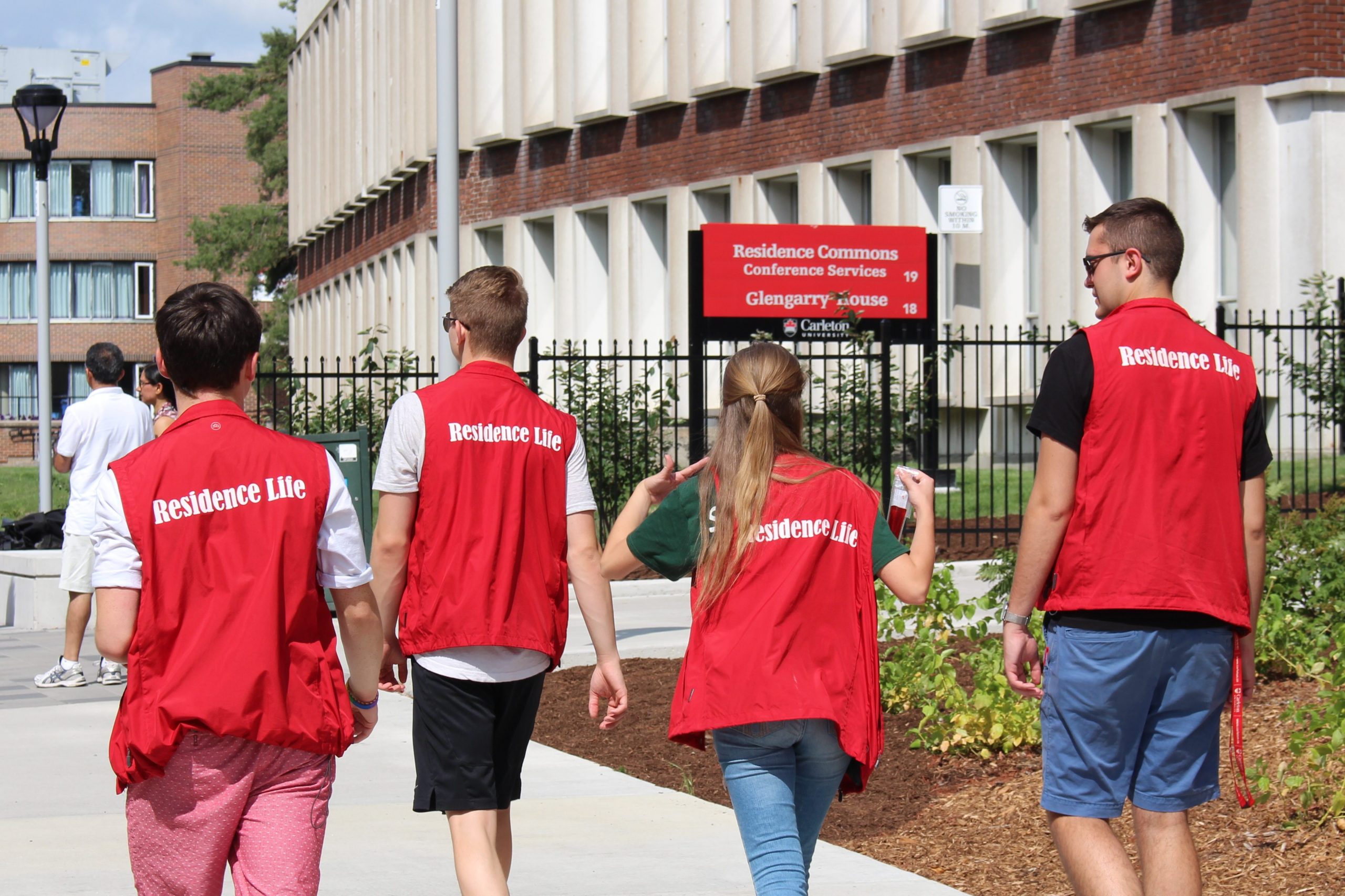 Four people wearing red Residence Life vests walk in the Residence quad