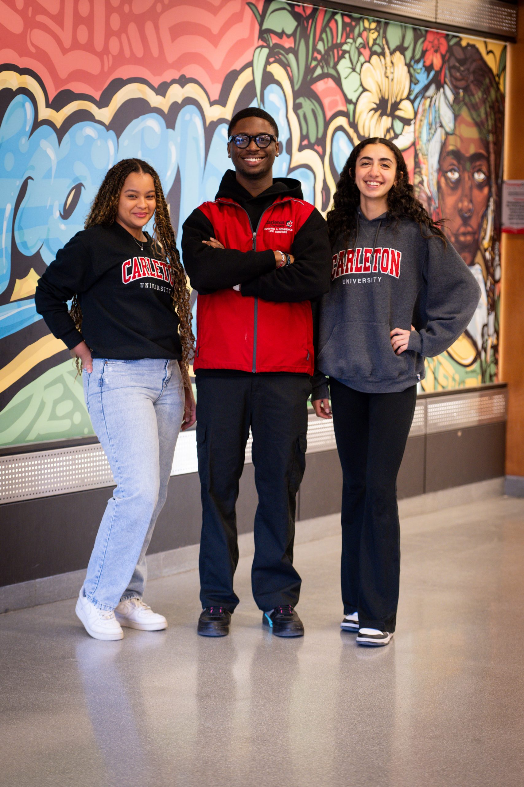 Three Carleton students smiling in front of the Teraanga Common mural.