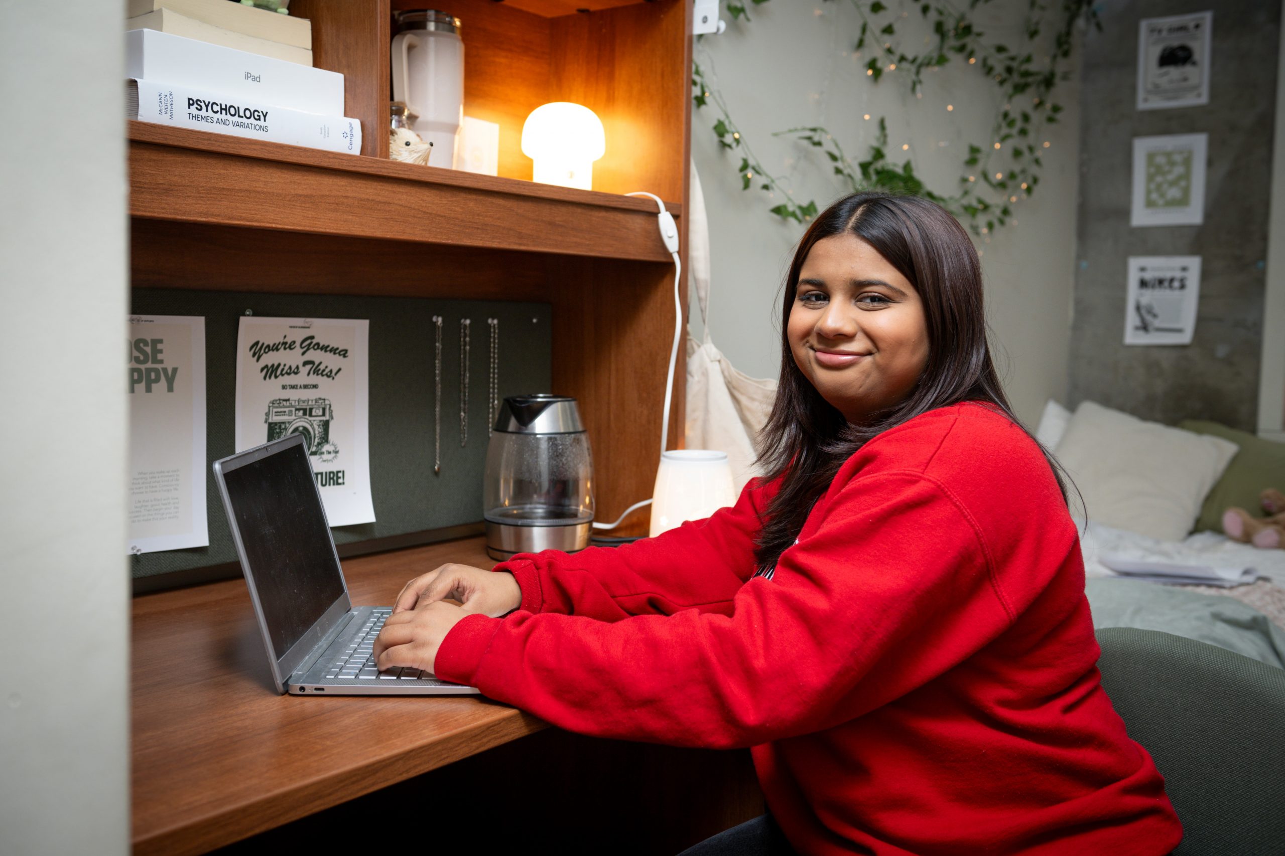 A Carleton student in a dorm room types on a laptop at a wooden desk, surrounded by plants, books, and a softly lit lamp.