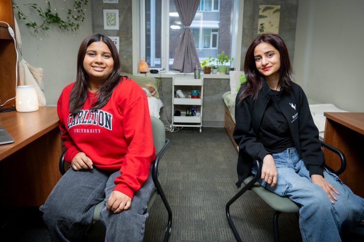 Two Carleton students sit in a cozy room decorated with plants, two desk workstations, and a large window allowing natural light.