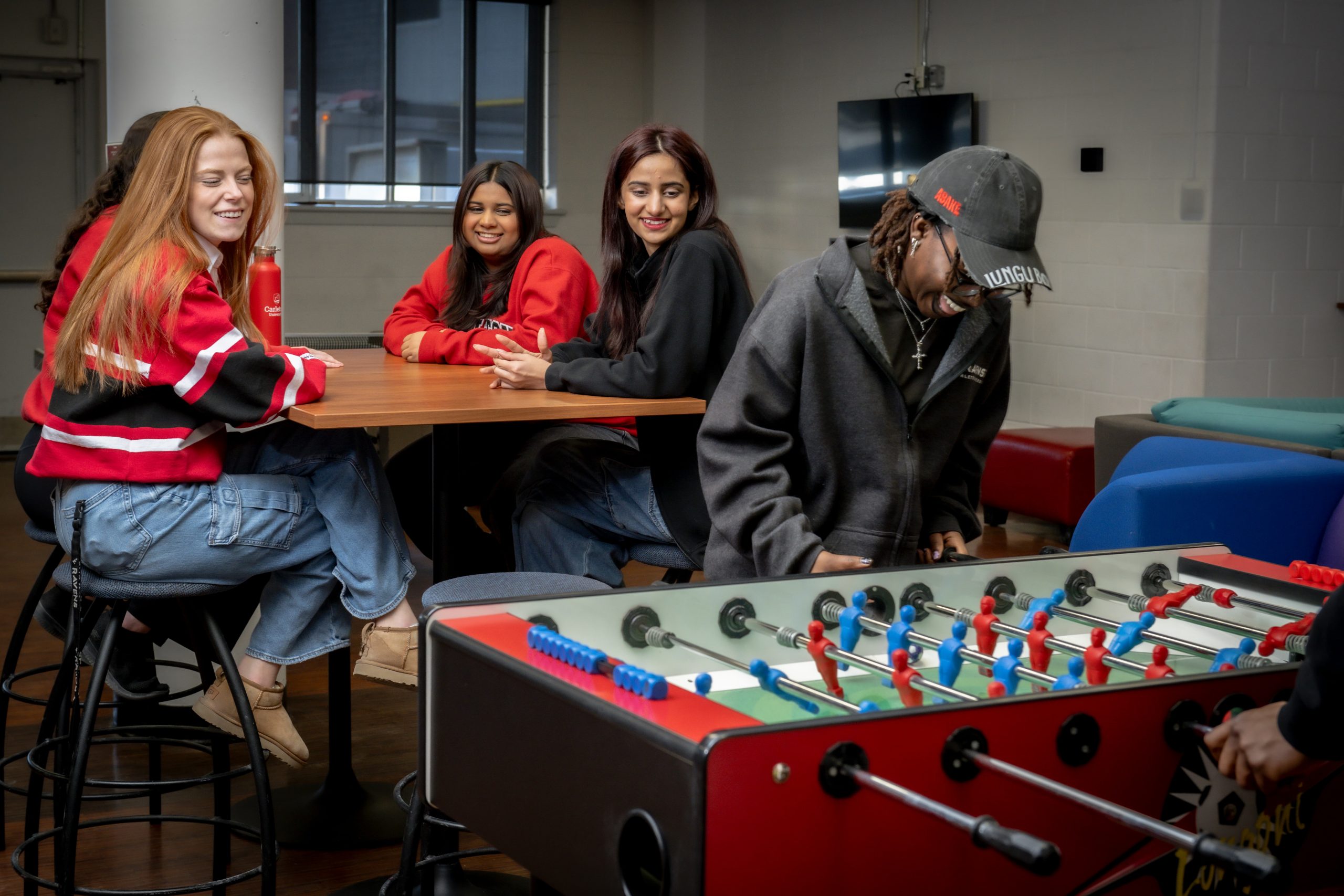 A group of Carleton students sit at a table watching others play foosball in Ravens Roost.