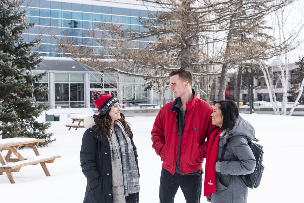 Three students engage in conversation outdoors on a snowy day, surrounded by snow-covered trees and the Carleton library in the back