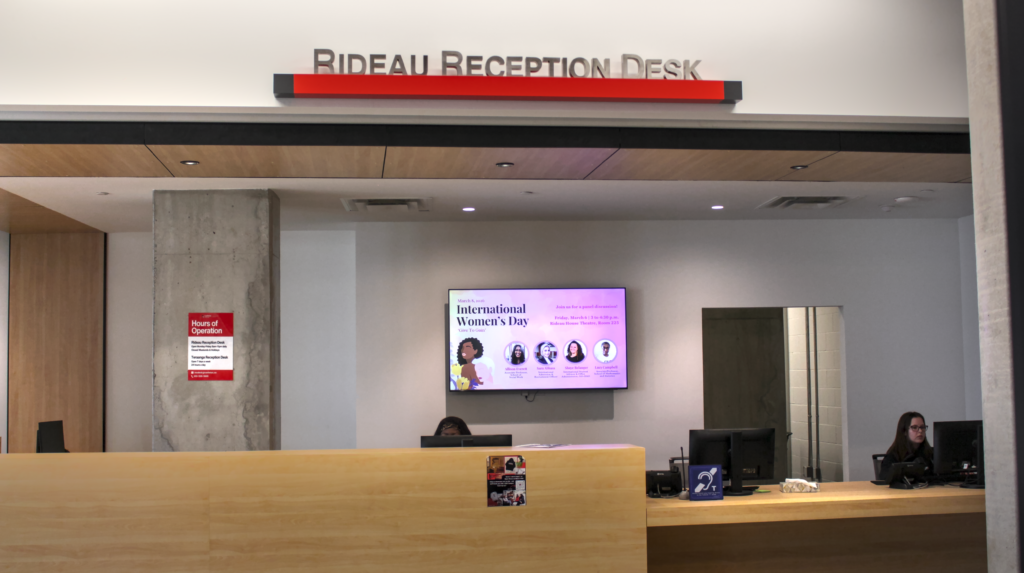 A head on shot of the Rideau Reception desk, with the sign above the two workers in front of their computers behind the desk, and a large on the wall TV, in the back, showcasing on-going events on campus