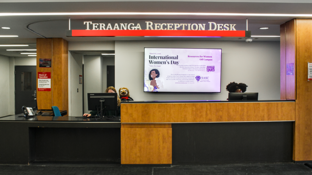 A head on shot of the Teraanga Commons Reception Desk with the sign above the 2 workers who are behind their computers at the desk, with a large TV behind them displaying on-going events on campus